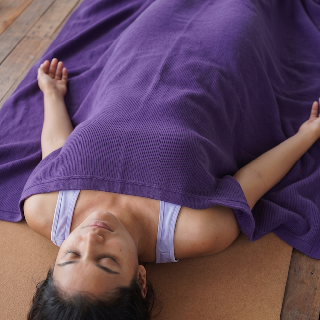 Woman resting in savasana covered by a purple cotton yoga blanket